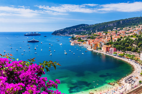 image of the french riviera coastline, blue mediterranean sea and buildings lining the coast line. there are purple flowers, mountains, boats in the water and white sand.