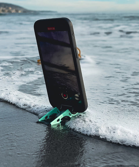 An iPhone on a Pocket Tripod on a the shore with a tide coming in, showing the Pocket Tripod elevates the phone to protect ports from water or anything on the surface